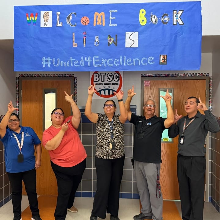 Five people pointing to a blue sign that says "Welcome Back Lions."